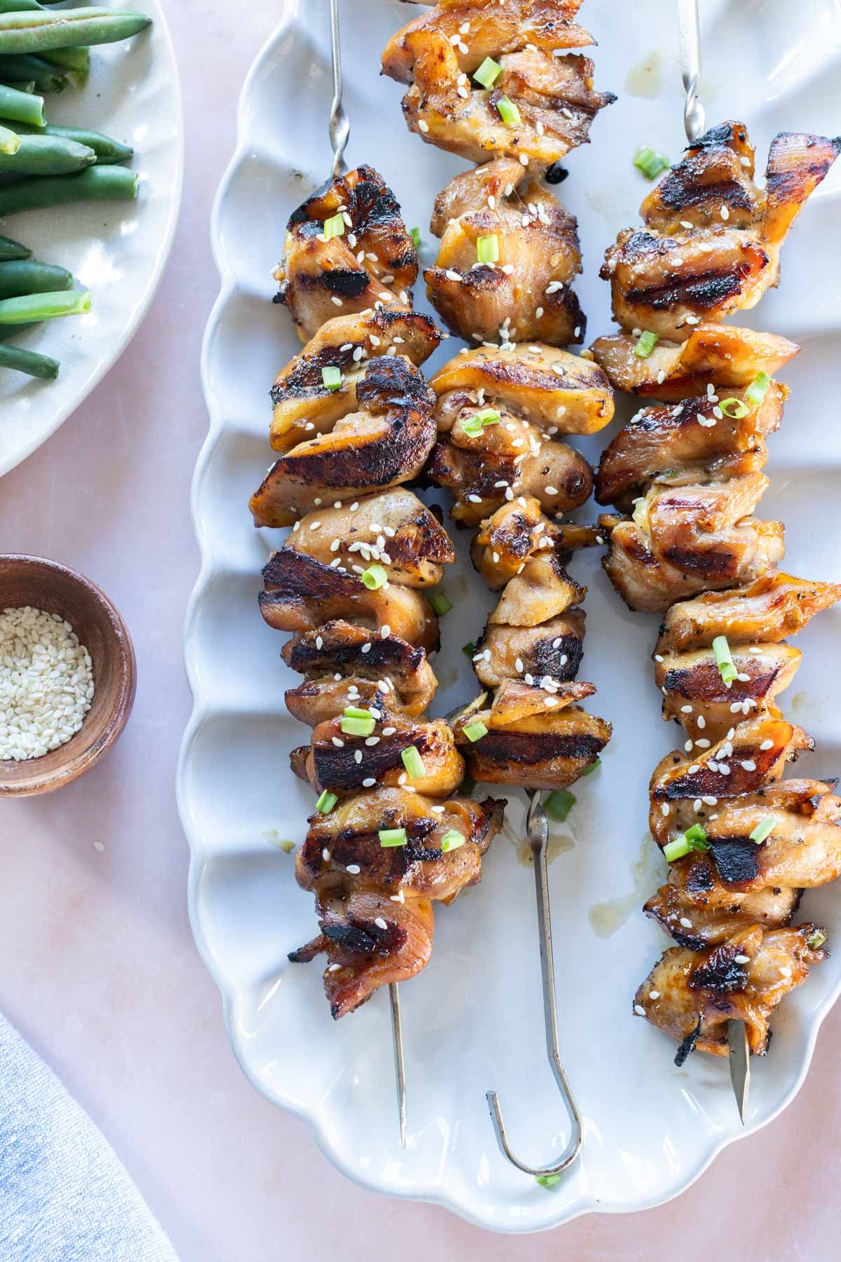 Three grilled chicken skewers garnished with sesame seeds and chopped green onions on a white plate, with a bowl of sesame seeds and green beans nearby.