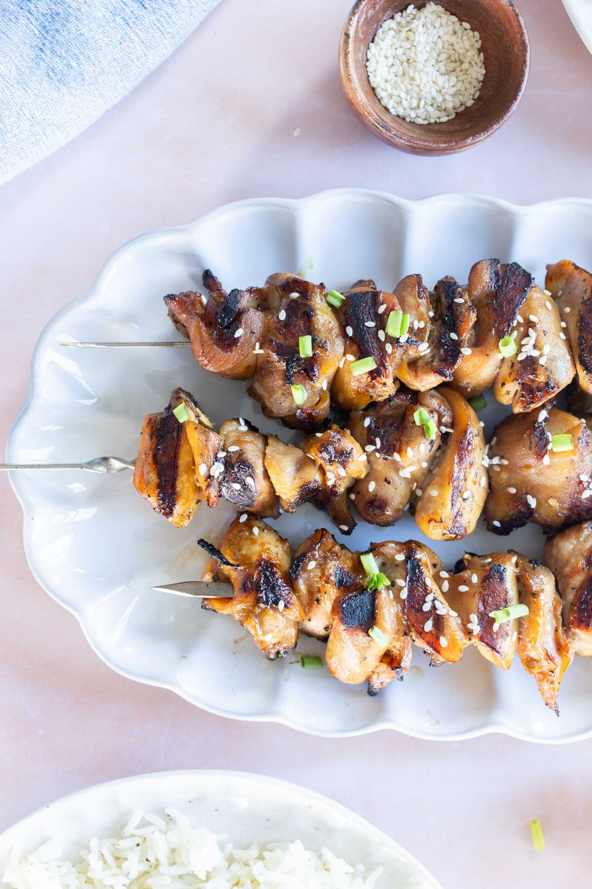Grilled chicken skewers topped with chopped green onions and sesame seeds are served on a white plate next to a bowl of sesame seeds and a partial view of white rice.