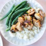 A plate with grilled chicken pieces on white rice, steamed green beans on the side, with a small bowl of sesame seeds nearby.