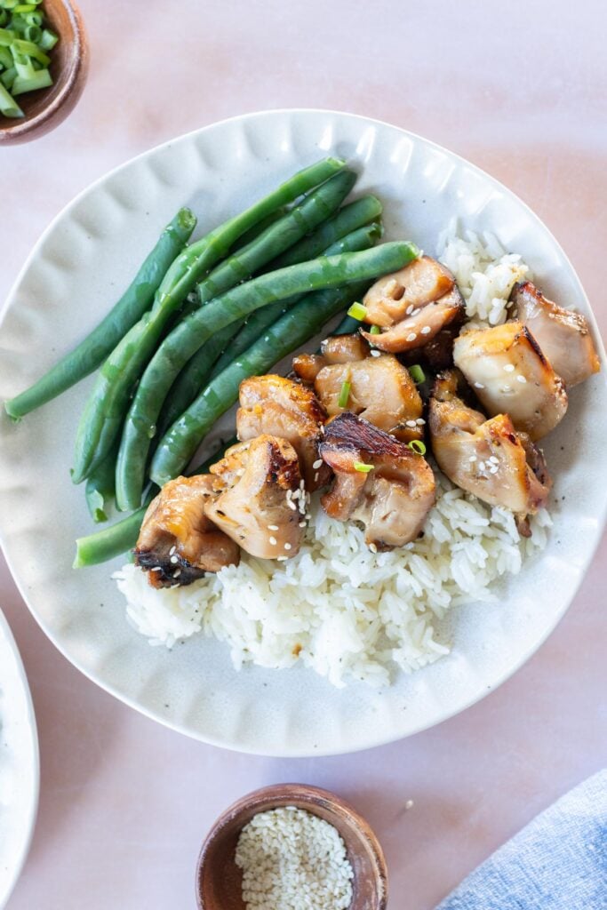 A plate with grilled chicken pieces on white rice, steamed green beans on the side, with a small bowl of sesame seeds nearby.