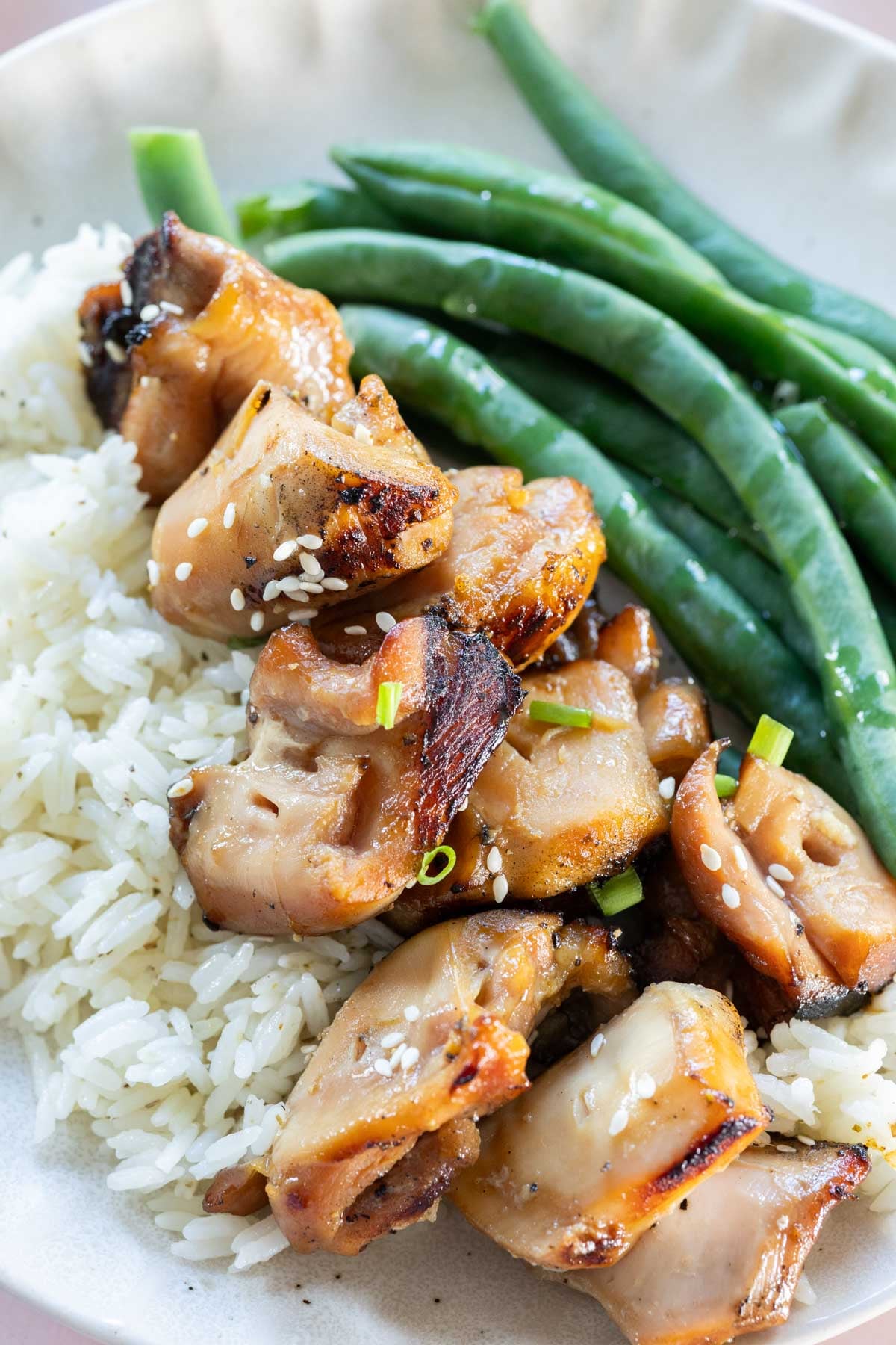 Plate of grilled teriyaki chicken pieces garnished with sesame seeds served over white rice, and steamed green beans.