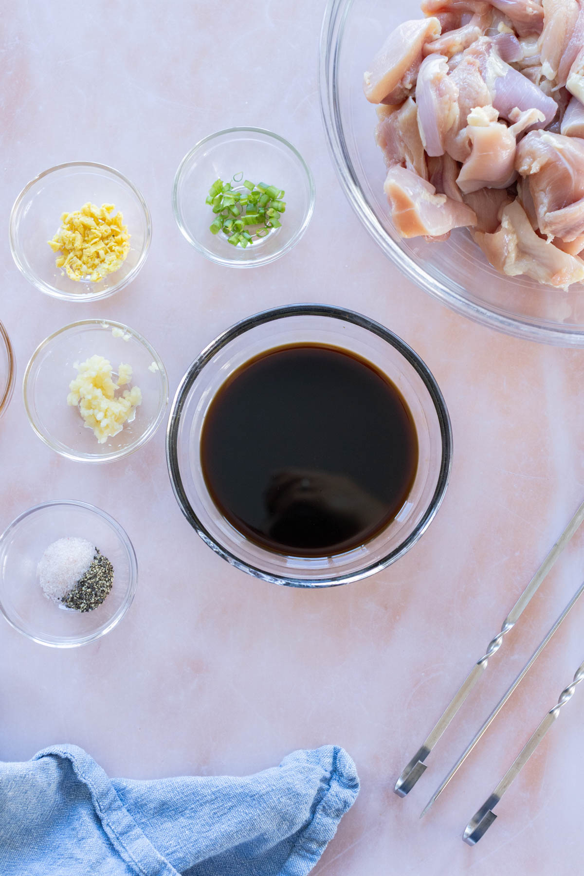 A bowl of chopped raw chicken, small bowls of soy sauce, chopped green onion, minced ginger, minced garlic, salt and pepper on a light surface with metal tongs and a blue cloth.