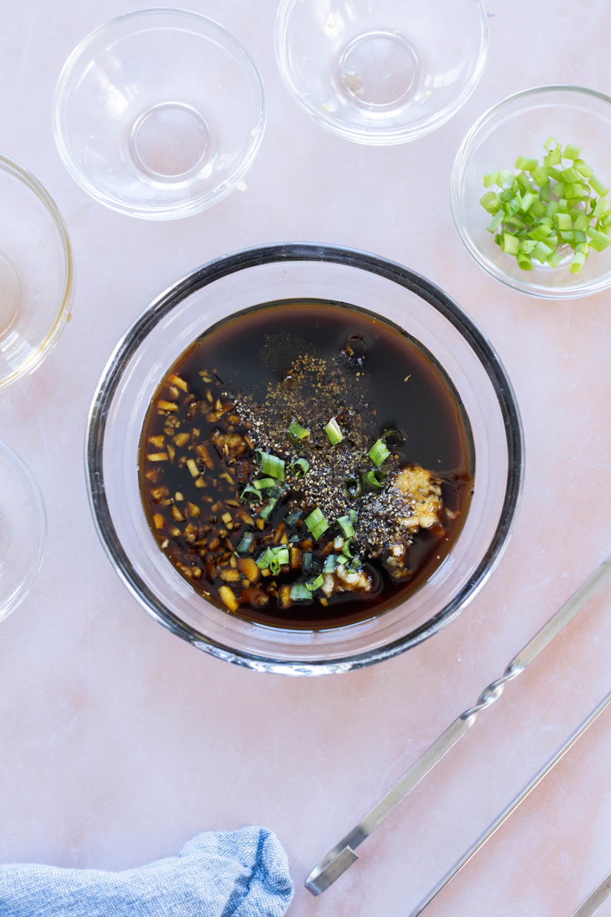 A glass bowl filled with a teriyaki marinade, surrounded by empty glass bowls and metal tongs on a light surface.