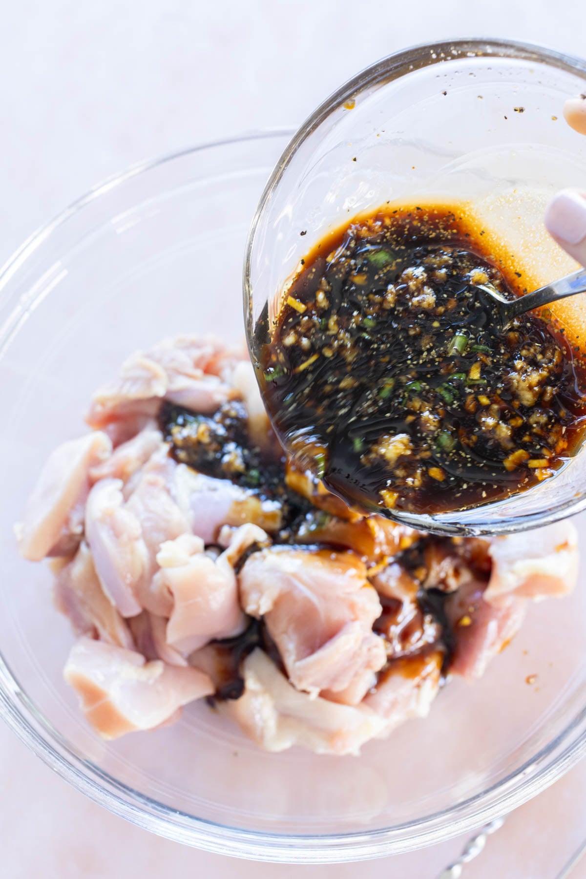 A hand pours a bowl of dark marinade with spices over raw pieces of chicken in a clear glass mixing bowl.