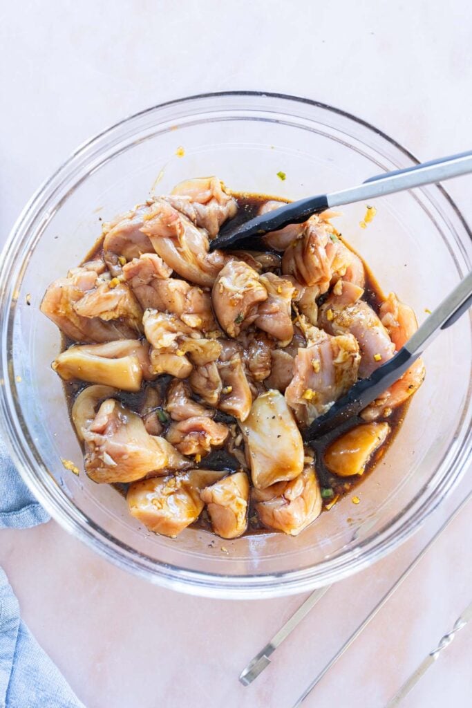 Raw chicken pieces marinating in a glass bowl with brown sauce, next to a pair of metal tongs on a light surface.