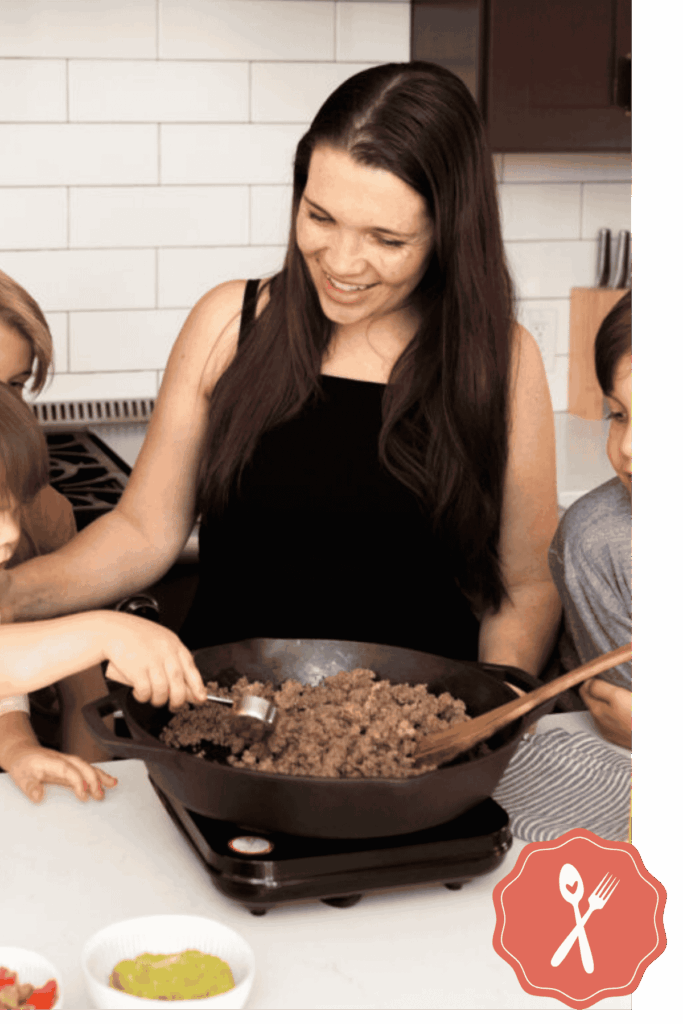 A woman stands at a kitchen counter, smiling as children help cook ground meat in a cast iron skillet. Small bowls with ingredients are nearby.
