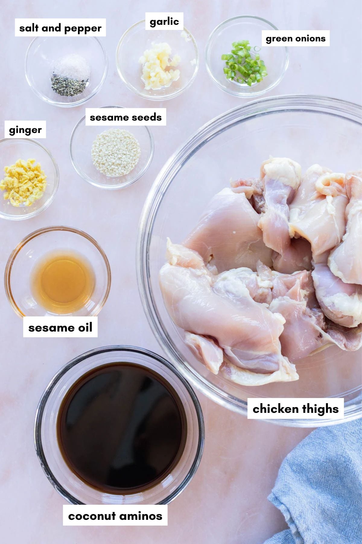 Labeled bowls containing chicken thighs, coconut aminos, sesame oil, sesame seeds, salt and pepper, garlic, ginger, and chopped green onions, all arranged on a counter.