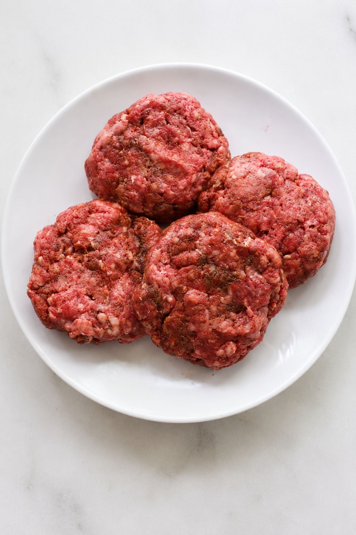 Four raw ground beef burger patties arranged on a white plate, viewed from above on a light-colored surface, ready to be transformed into flavorful Hawaiian Burgers topped with grilled pineapple.