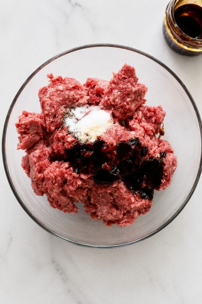 Ground beef in a glass bowl topped with dark sauce and seasonings, reminiscent of the flavors found in Hawaiian Burgers, with a small glass container of additional sauce beside it on a white surface. The sweet and savory profile is perfect for pairing with grilled pineapple for an added tropical twist.