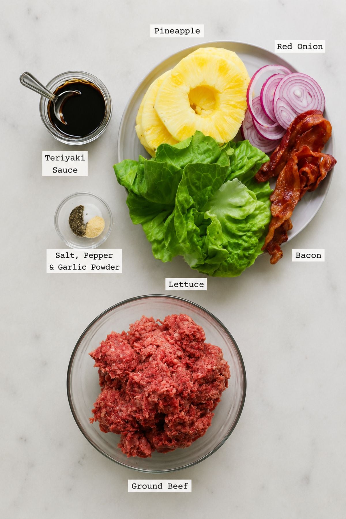 A plate with grilled pineapple, red onion, bacon, and lettuce sits next to bowls of teriyaki sauce, seasonings, and ground beef, all labeled on a white surface.