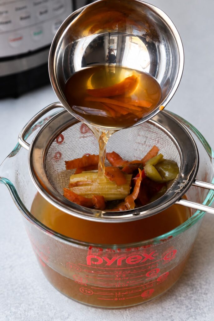 Instant Pot vegetable broth is being poured through a metal strainer with vegetables into a glass measuring cup on a countertop.