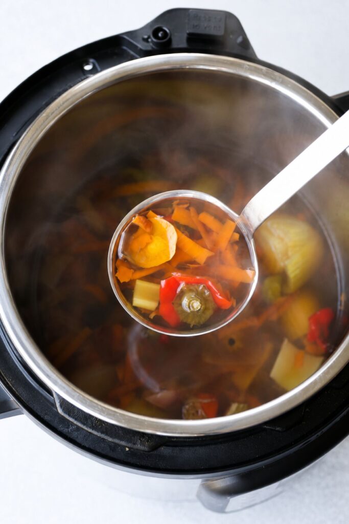A ladle holds instant pot vegetable broth with carrots, peppers, and onion above an open electric pressure cooker filled with steaming broth.