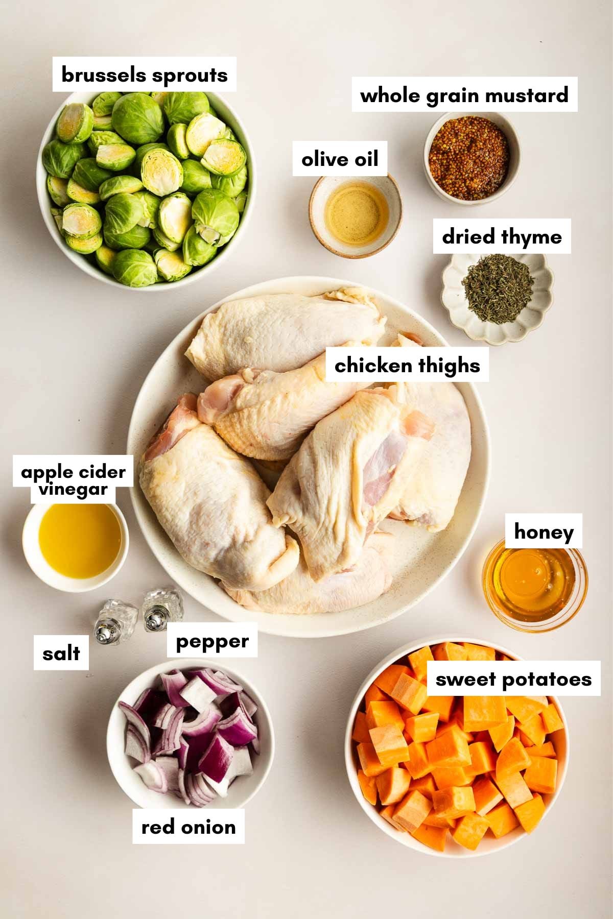 A flat lay of labeled ingredients for sheet pan chicken thighs, including chicken thighs, sweet potatoes, brussels sprouts, red onion, whole grain mustard, olive oil, dried thyme, apple cider vinegar, honey, salt, and pepper.