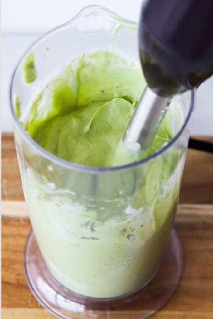 A hand blender is mixing a thick, green avocado mixture in a transparent plastic container on a wooden surface.