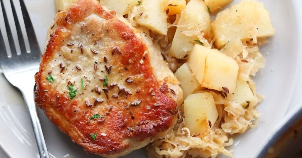 A cooked pork chop garnished with herbs sits on a plate next to cubed potatoes and sauerkraut, with a fork beside the food.