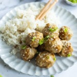 A white plate with rice and eight sesame-topped meatballs garnished with chopped green onions, with chopsticks resting on the plate.