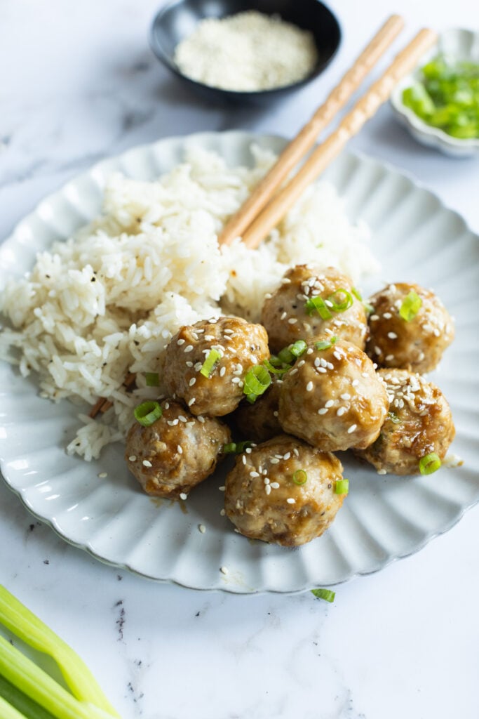 A white plate with rice and eight sesame-topped meatballs garnished with chopped green onions, with chopsticks resting on the plate.