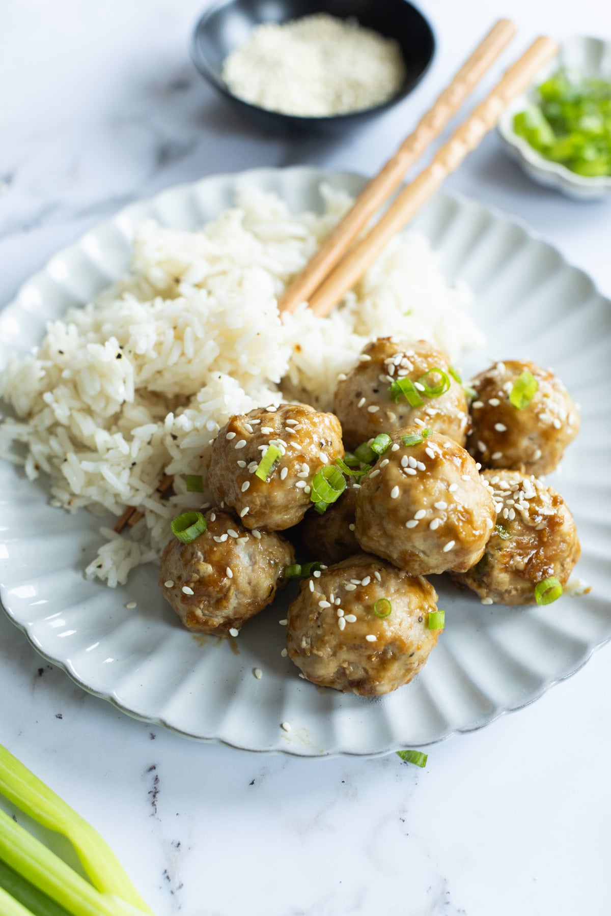 A white plate with rice and eight sesame-topped meatballs garnished with chopped green onions, with chopsticks resting on the plate.