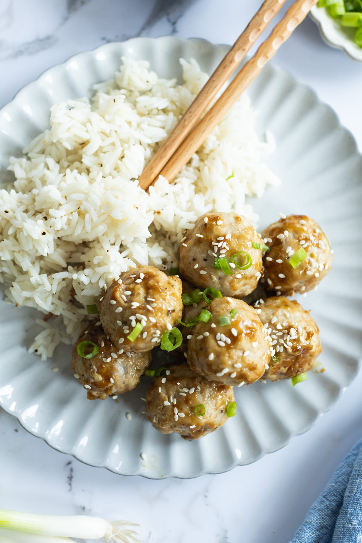 Plate with a serving of white rice and eight glazed meatballs, garnished with sliced green onions and sesame seeds, with wooden chopsticks resting on top.