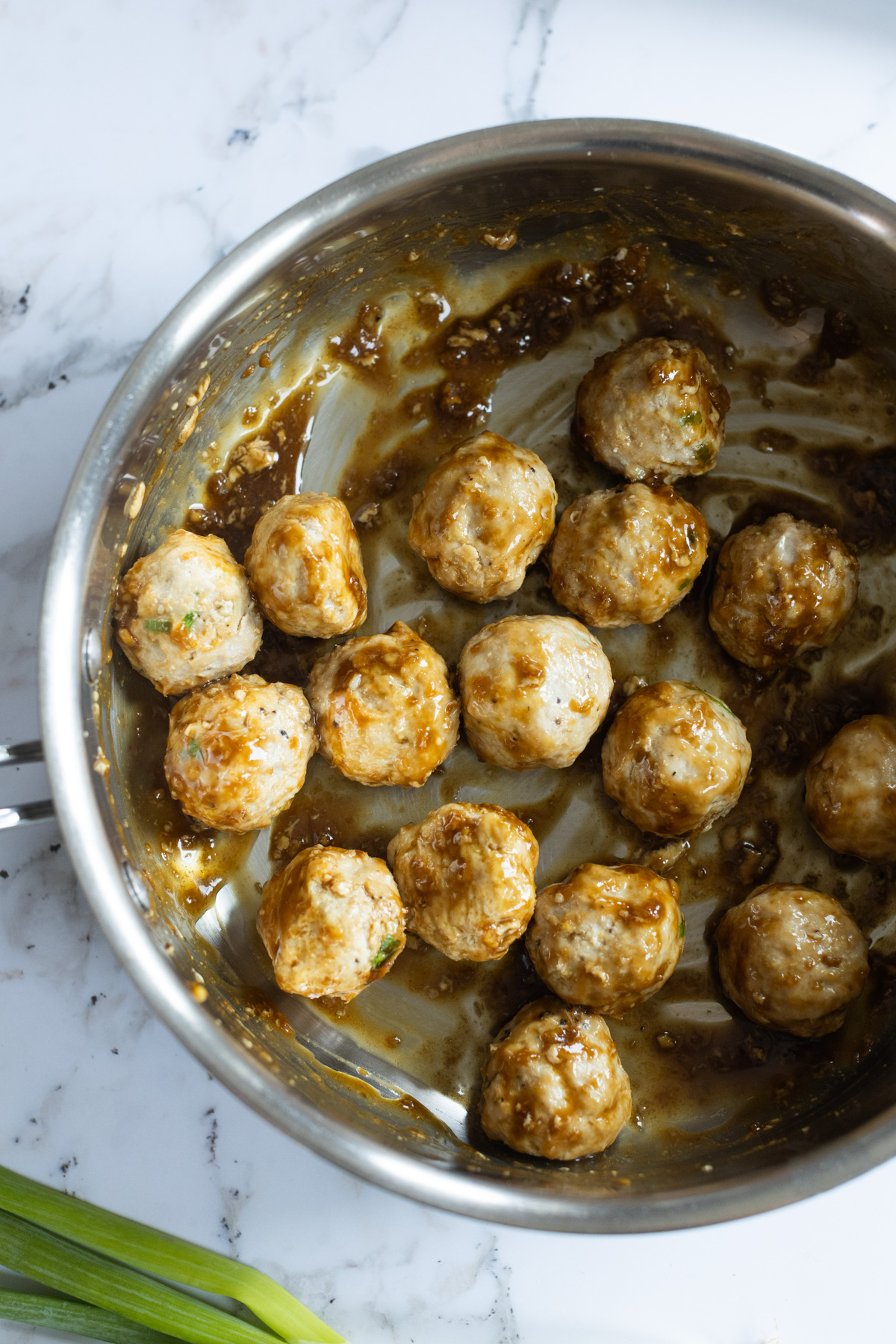 Glazed meatballs coated in sauce in a stainless steel skillet sits on a white marble surface.