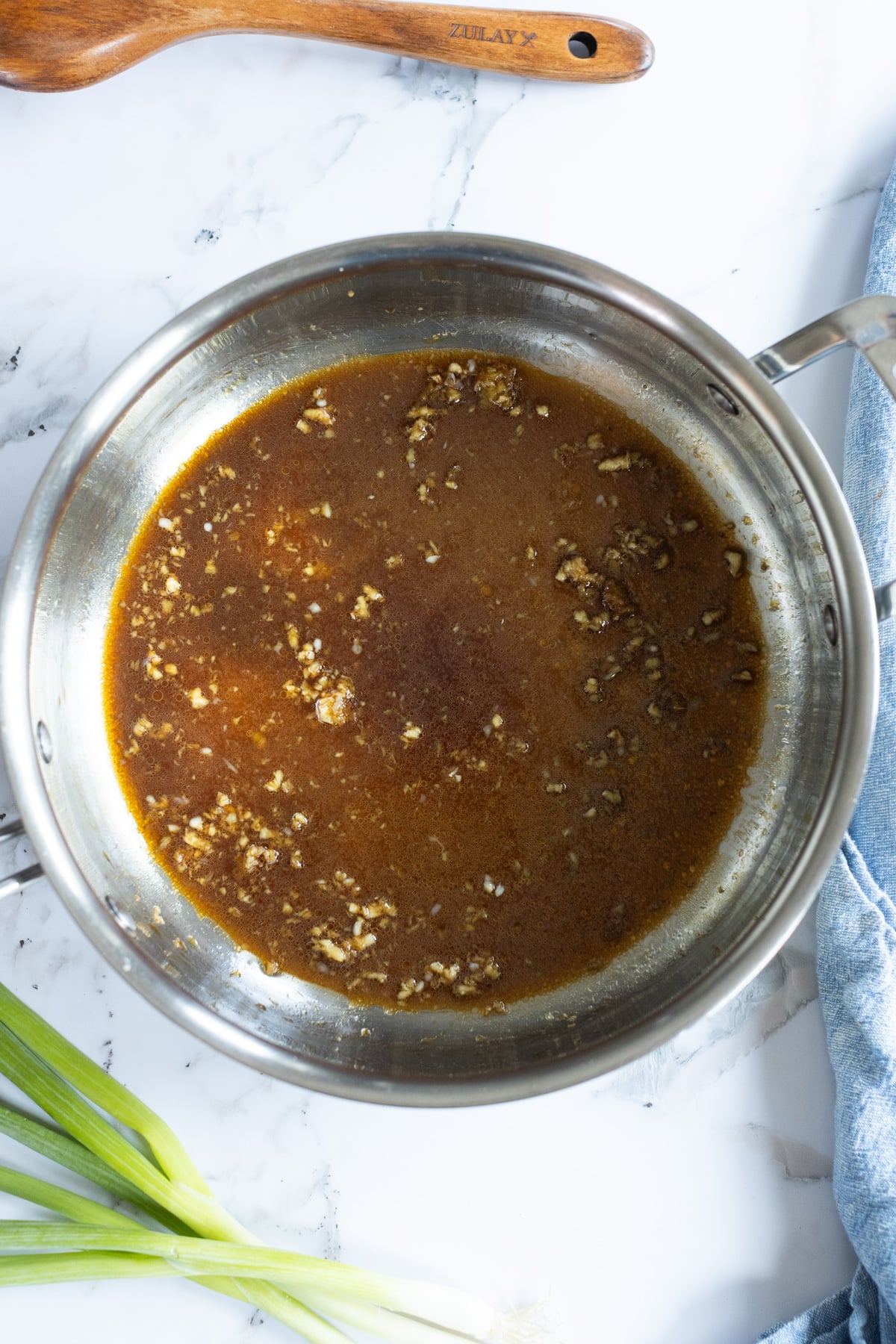 A stainless steel saucepan with brown sauce and minced garlic sits on a marble counter next to green onions and a blue cloth.