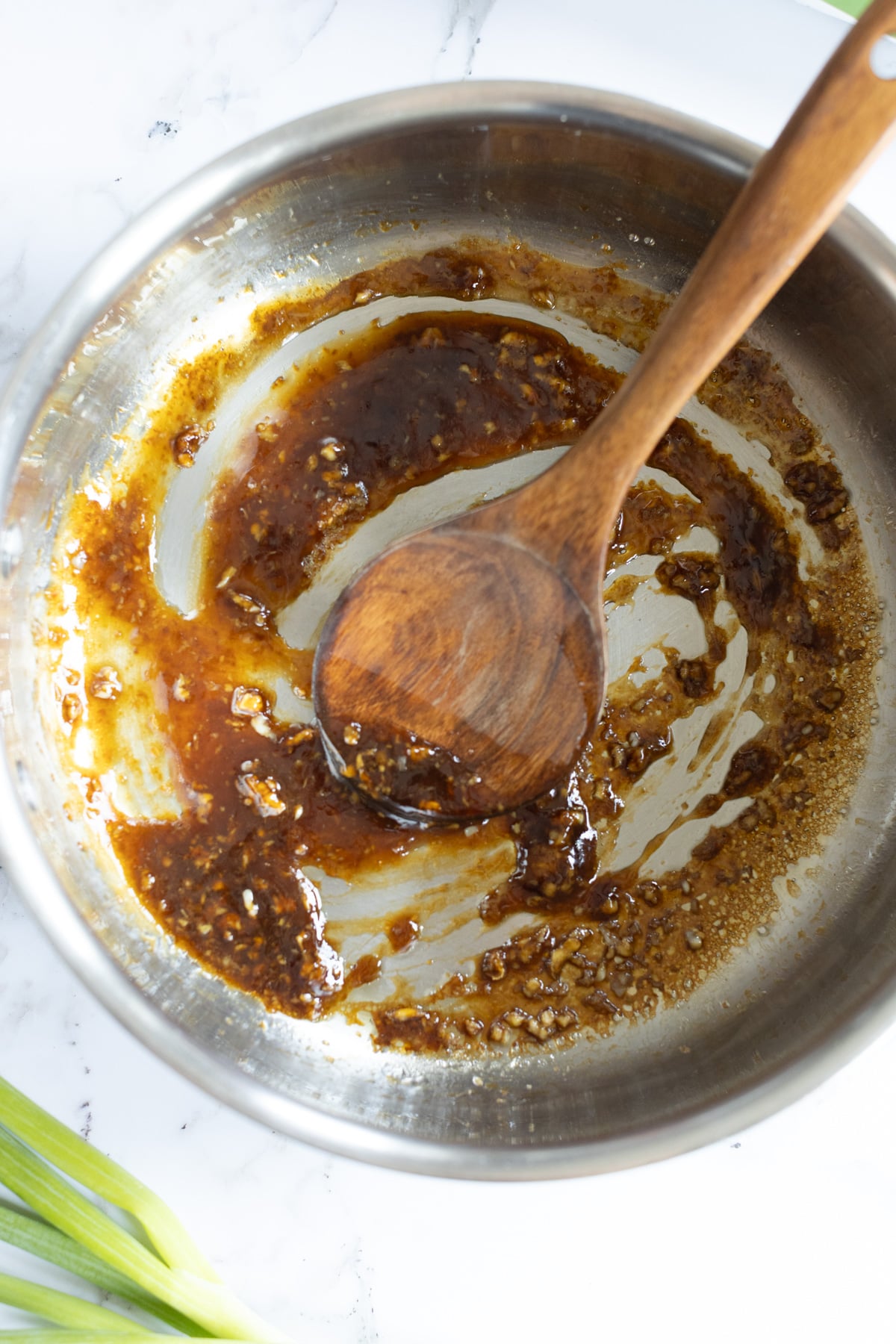 A metal bowl with a wooden spoon mixing a thick, dark brown sauce containing visible minced garlic or ginger, on a white surface with a green onion nearby.