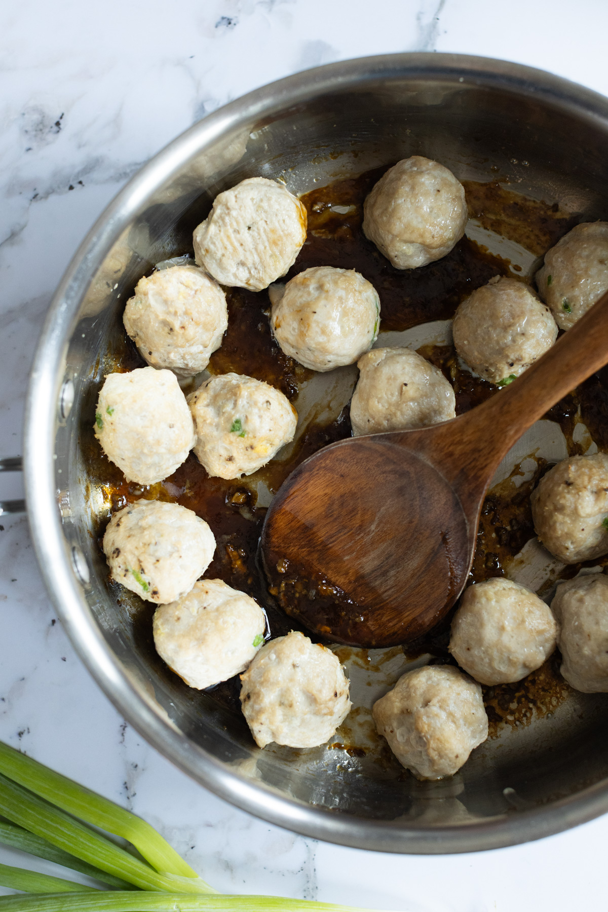 Chicken meatballs cooking in a stainless steel pan with brown sauce, a wooden spoon, and green onions visible on a marble countertop.