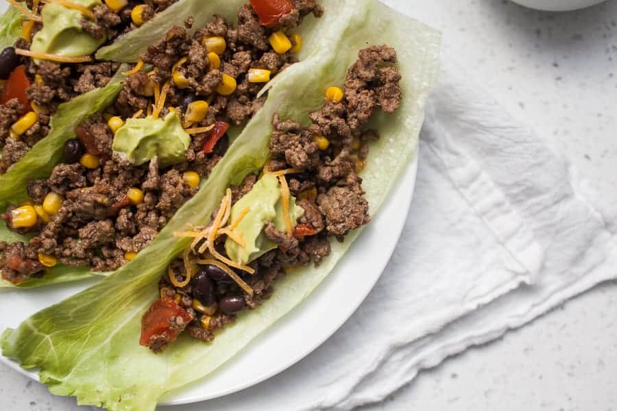 Ground beef, black beans, corn, diced tomatoes, avocado, and shredded cheese served on lettuce leaves on a white plate.