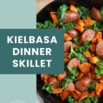 A cast iron skillet filled with sliced kielbasa, sweet potatoes, and kale sits on a wooden surface next to garlic cloves.