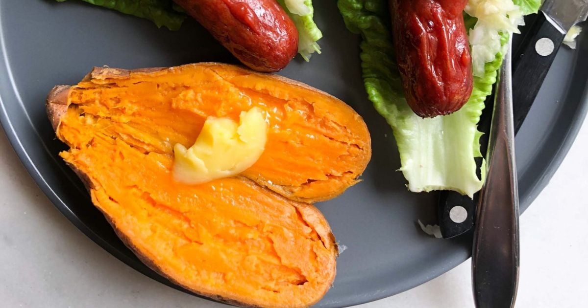 A microwaved sweet potato with a pat of butter sits on a plate next to lettuce leaves topped with sausage. A knife rests beside the food.