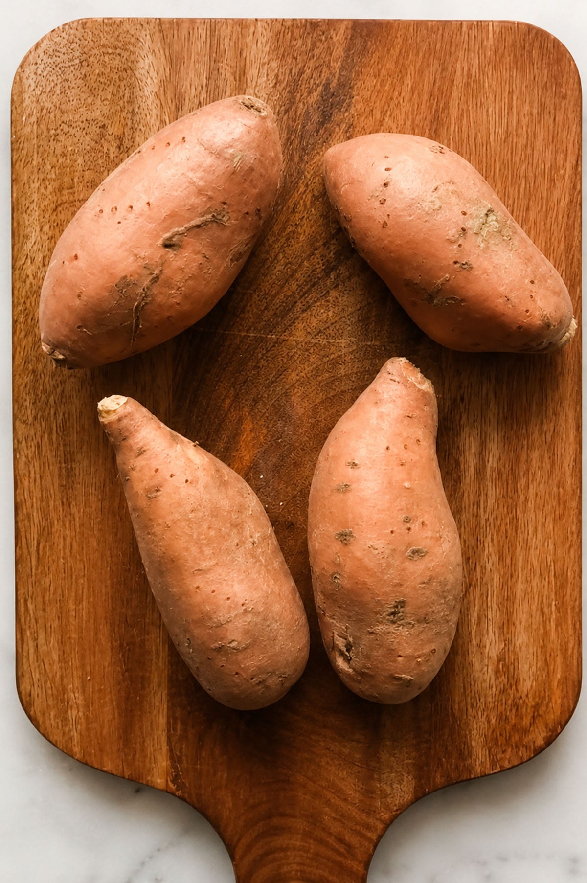 Four raw sweet potatoes on a wooden cutting board, arranged in a rough diamond shape.