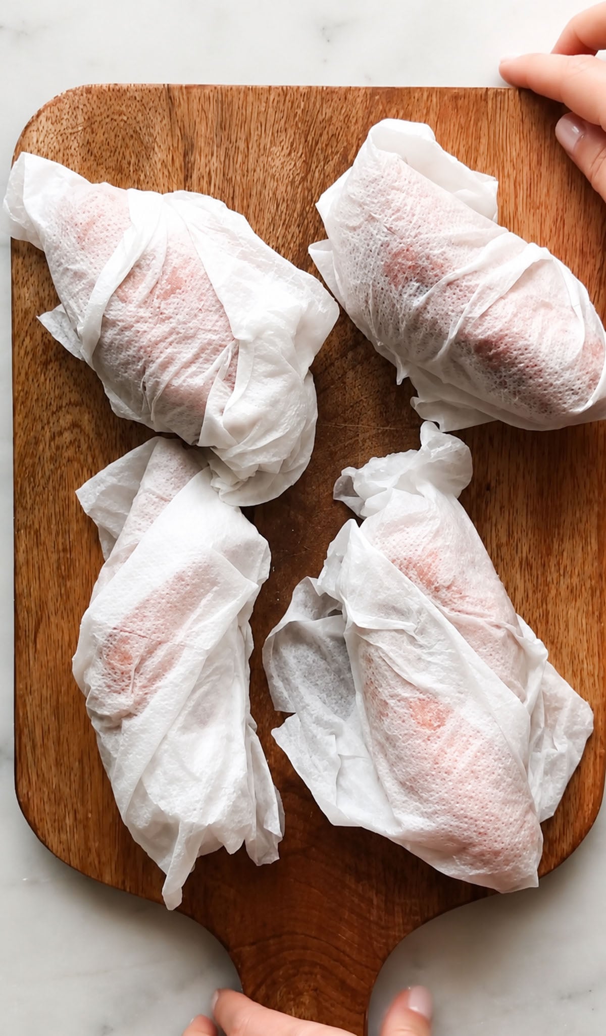 Four pieces of food wrapped in white paper towels are arranged on a wooden cutting board, with hands visible at the edges.