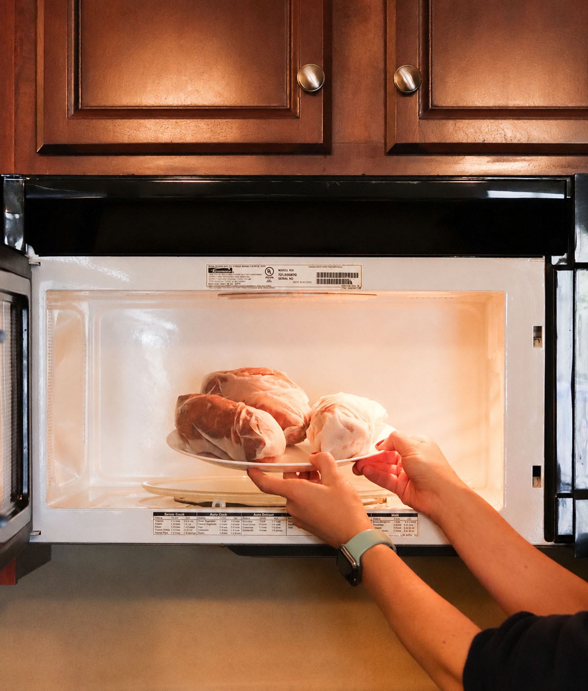 A person places a plate with three wrapped bread loaves into a microwave below brown cabinets.