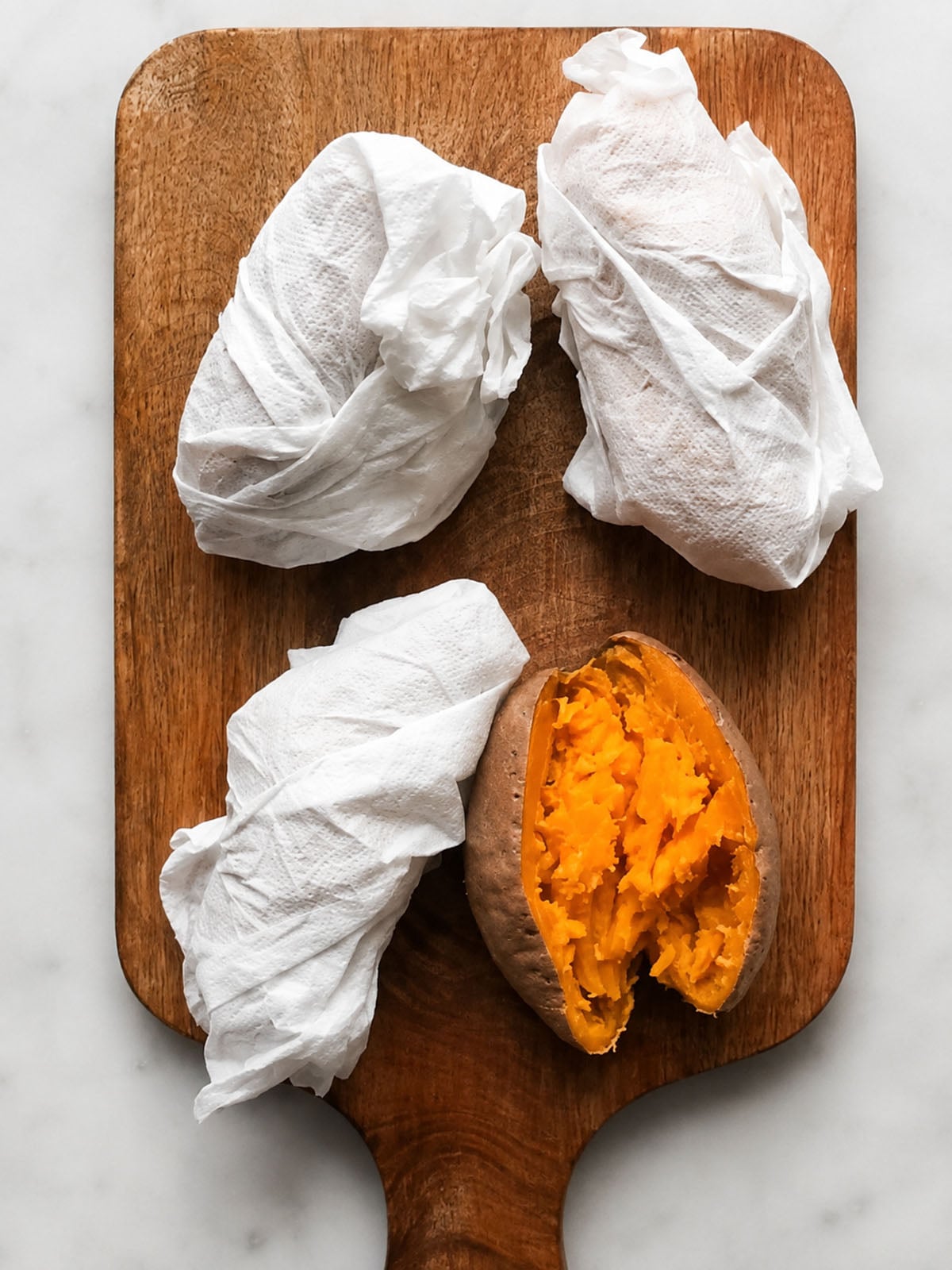 Three sweet potatoes wrapped in paper towels and one unwrapped, cooked sweet potato on a wooden cutting board.