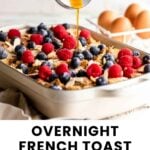 A pan of French toast casserole topped with berries, with syrup being poured over it; eggs and a spoon are in the background.