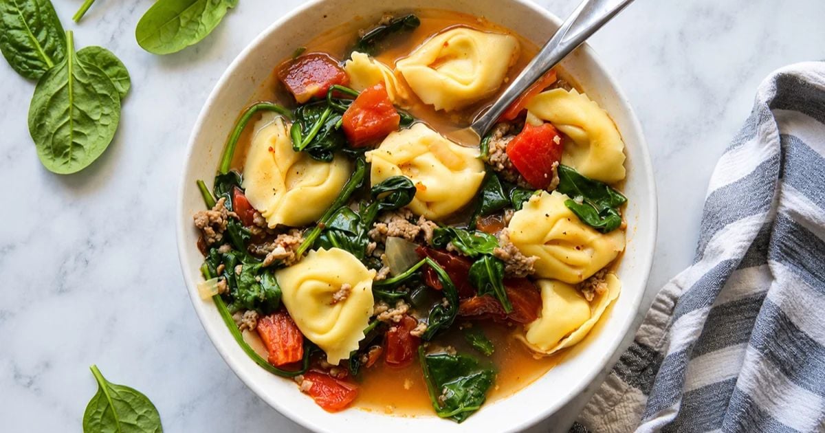 Bowl of soup with cheese tortellini, spinach, tomatoes, ground meat, and broth, served with a spoon. Fresh spinach leaves and a striped cloth are nearby on a marble surface.