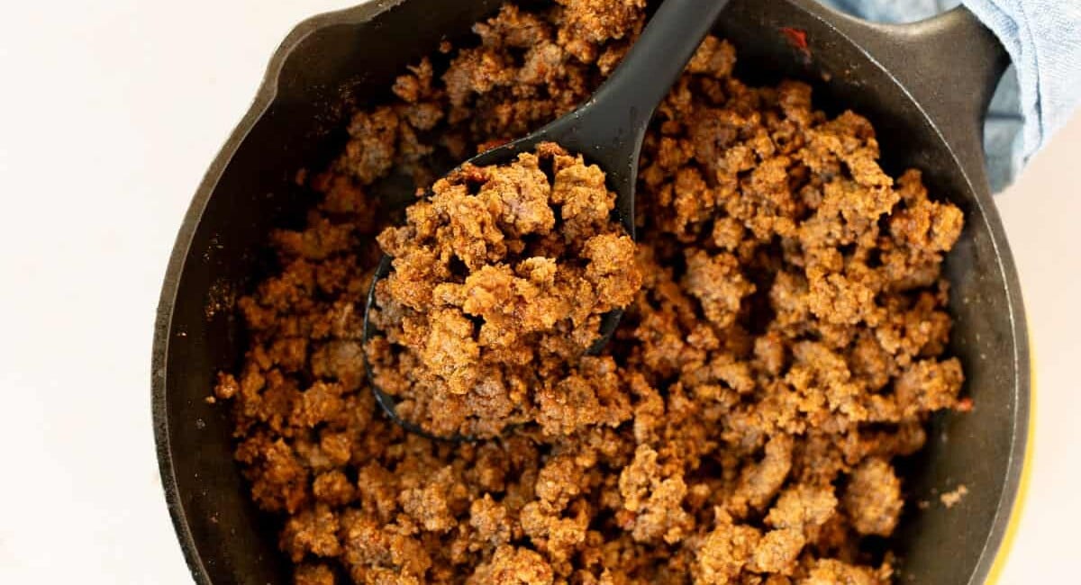 Cooked ground beef with seasoning in a black cast iron skillet, with a black spoon scooping some of the meat.
