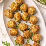 A white oval plate with fifteen cooked turkey meatballs garnished with chopped parsley, next to a bowl of green beans and a bowl of red sauce on a white surface.