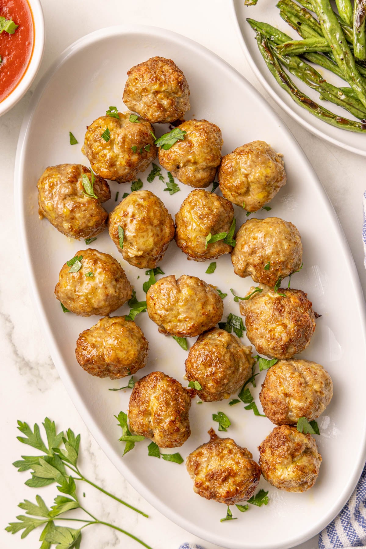 A white oval plate with fifteen cooked turkey meatballs garnished with chopped parsley, next to a bowl of green beans and a bowl of red sauce on a white surface.