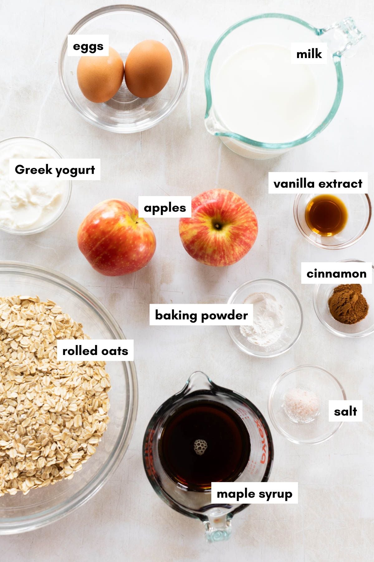 Ingredients in clear glass bowls with text overlay: rolled oats, apples, eggs, milk, yogurt, vanilla extract, cinnamon, salt, baking powder, and syrup.
