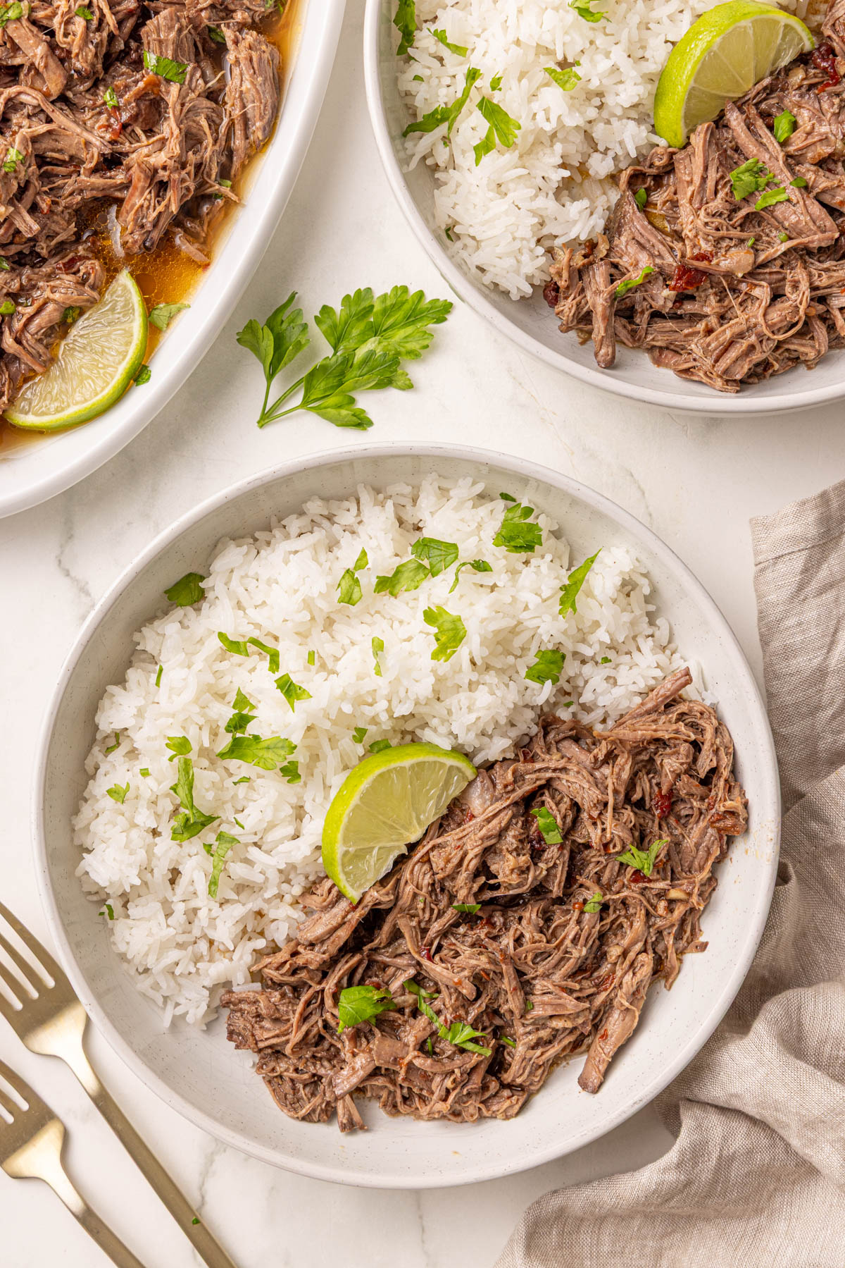 A bowl of shredded beef and white rice garnished with chopped parsley and a lime wedge, with a similar plate and a serving dish nearby.