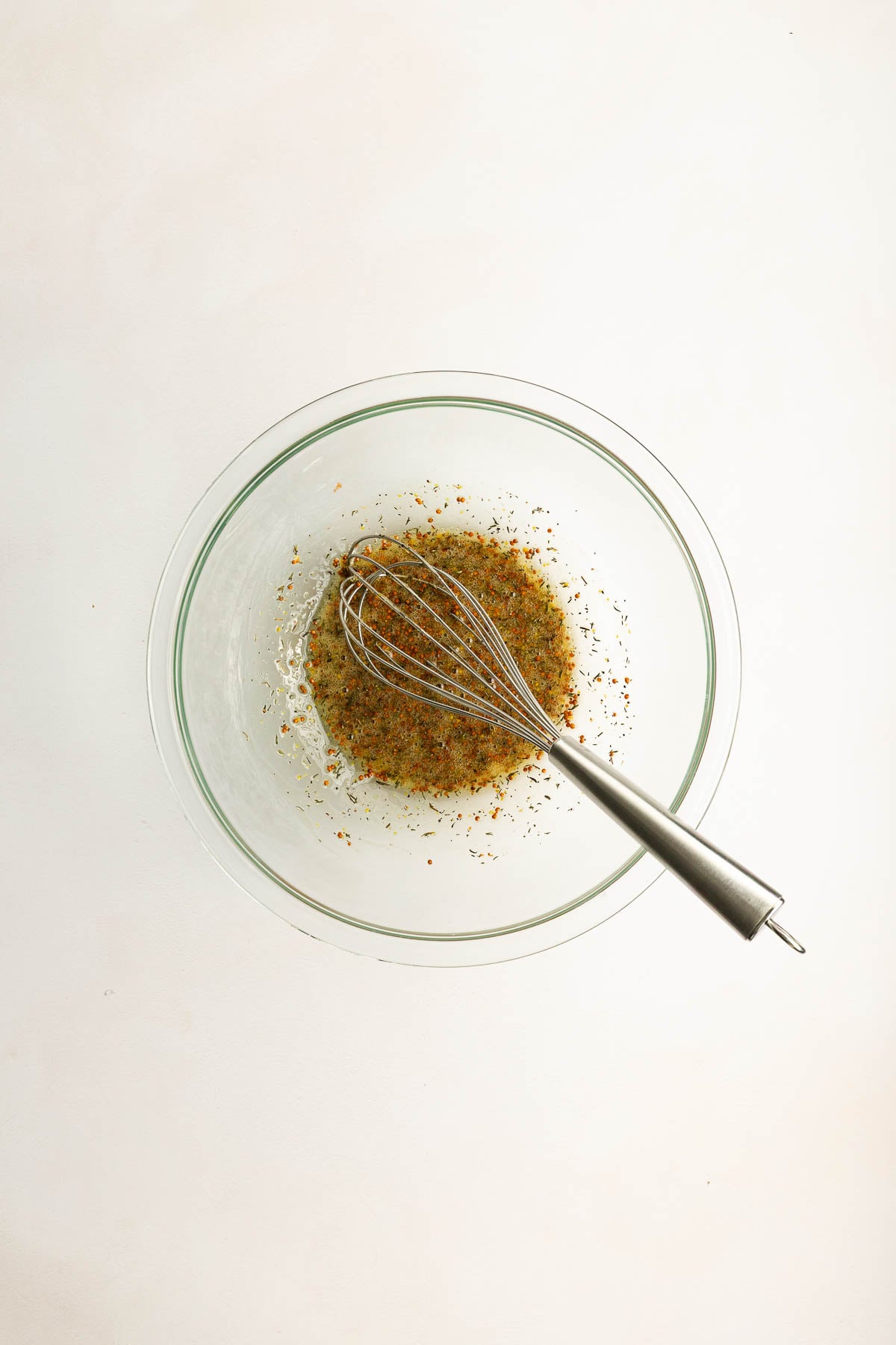 A metal whisk rests in a glass bowl containing a mixture of herbs and spices against a plain white background.