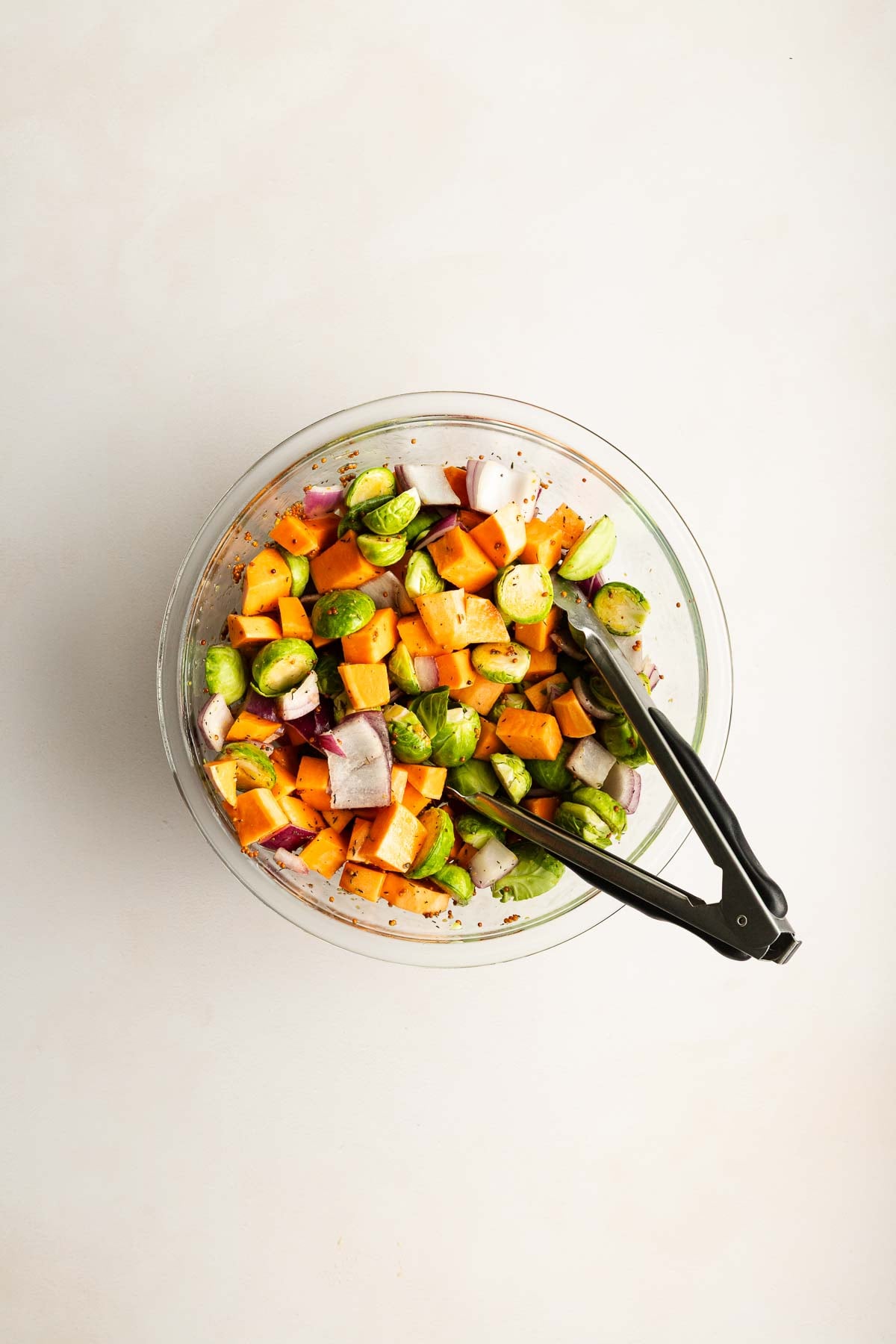 A glass bowl filled with chopped sweet potatoes, Brussels sprouts, and red onions with metal tongs resting on the side.