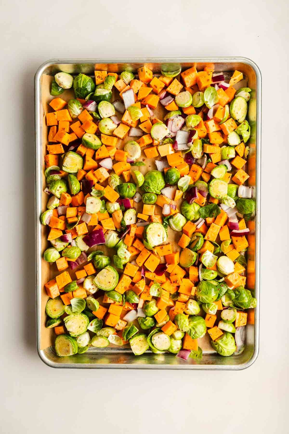 Chopped Brussels sprouts, sweet potatoes, and red onions on a large baking sheet, seasoned and ready to roast.