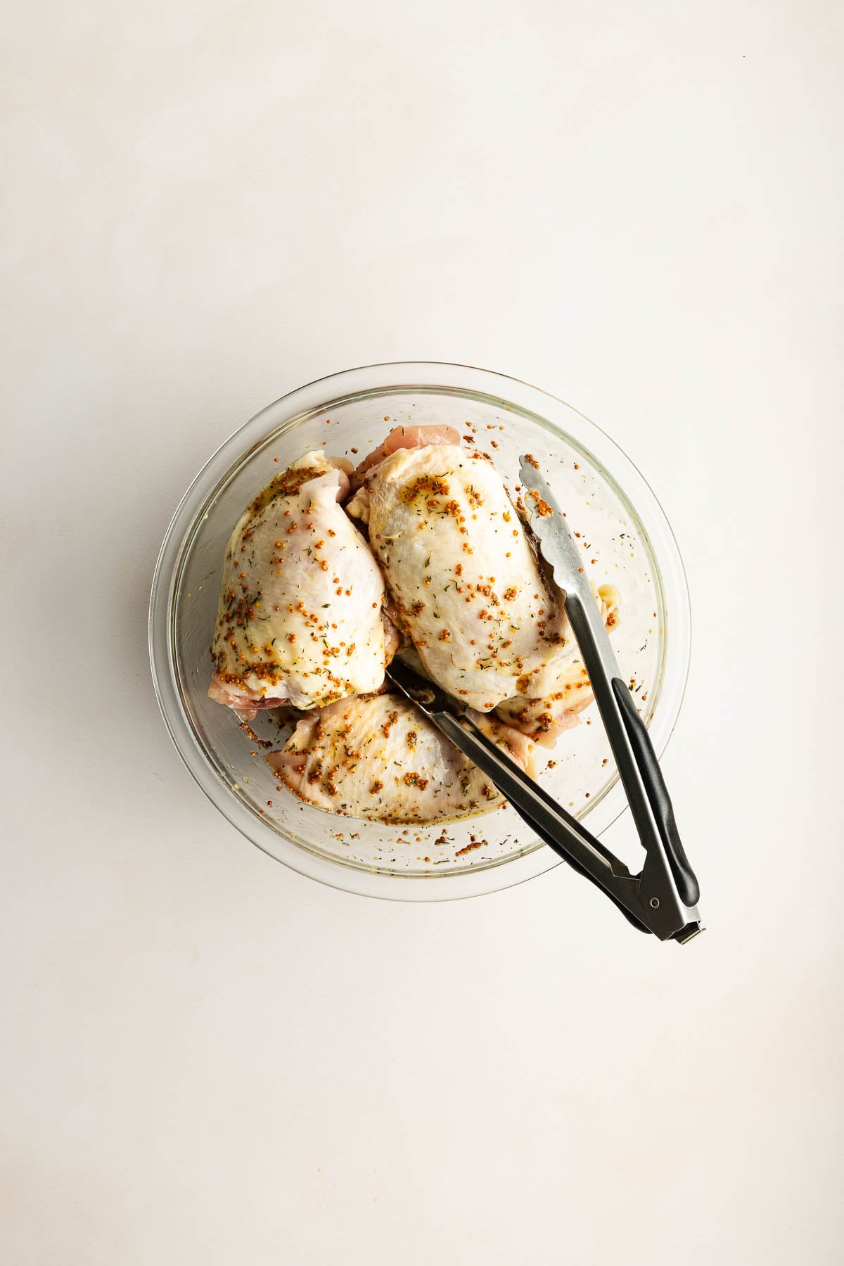 A glass bowl with four raw, seasoned chicken thighs and metal tongs on a white surface.