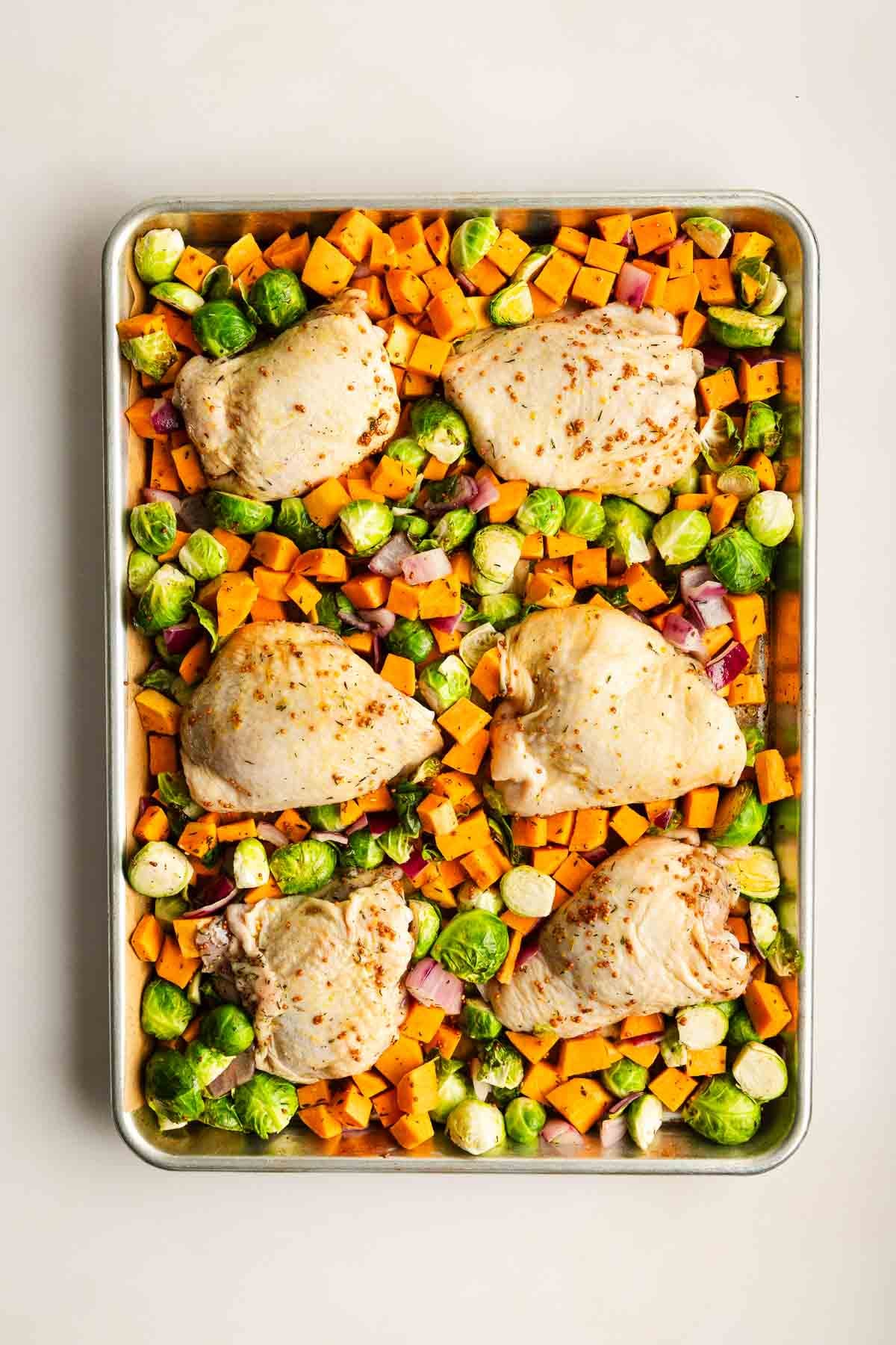 Sheet pan with five raw chicken thighs, diced sweet potatoes, Brussels sprouts, and chopped red onions, ready for roasting.