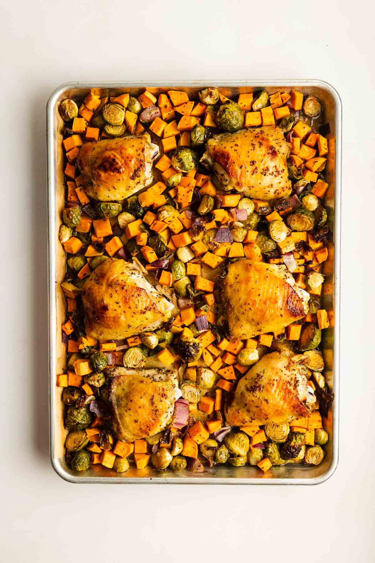 Sheet pan with six baked chicken thighs surrounded by roasted sweet potatoes, Brussels sprouts, and red onions on a white background.