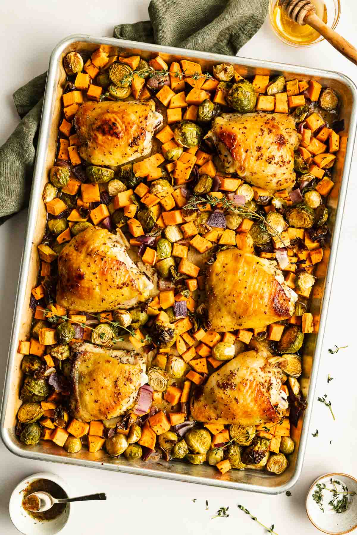 Sheet pan with baked chicken thighs, sweet potatoes, Brussels sprouts, and red onions, garnished with herbs.