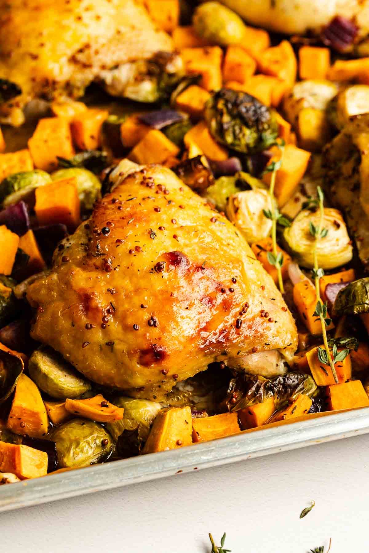 Roasted chicken thigh on a baking sheet with chopped sweet potatoes, brussels sprouts, red onions, and fresh thyme.
