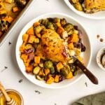 A plate of roasted chicken thigh served with Brussels sprouts, sweet potatoes, and red onions. A sheet pan with more roasted vegetables and a bowl of honey are visible nearby.